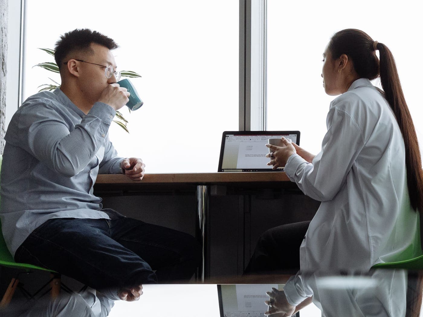 Two people seated at a table by a large window with a laptop, reflected in the glossy table top
