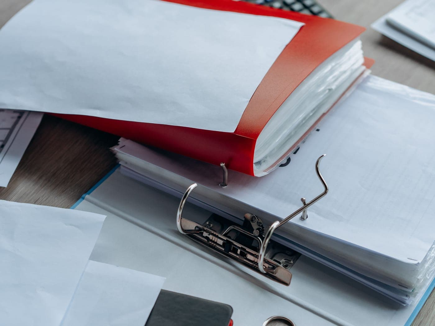 An open ring binder under a red folder and stacks of paper on a desk