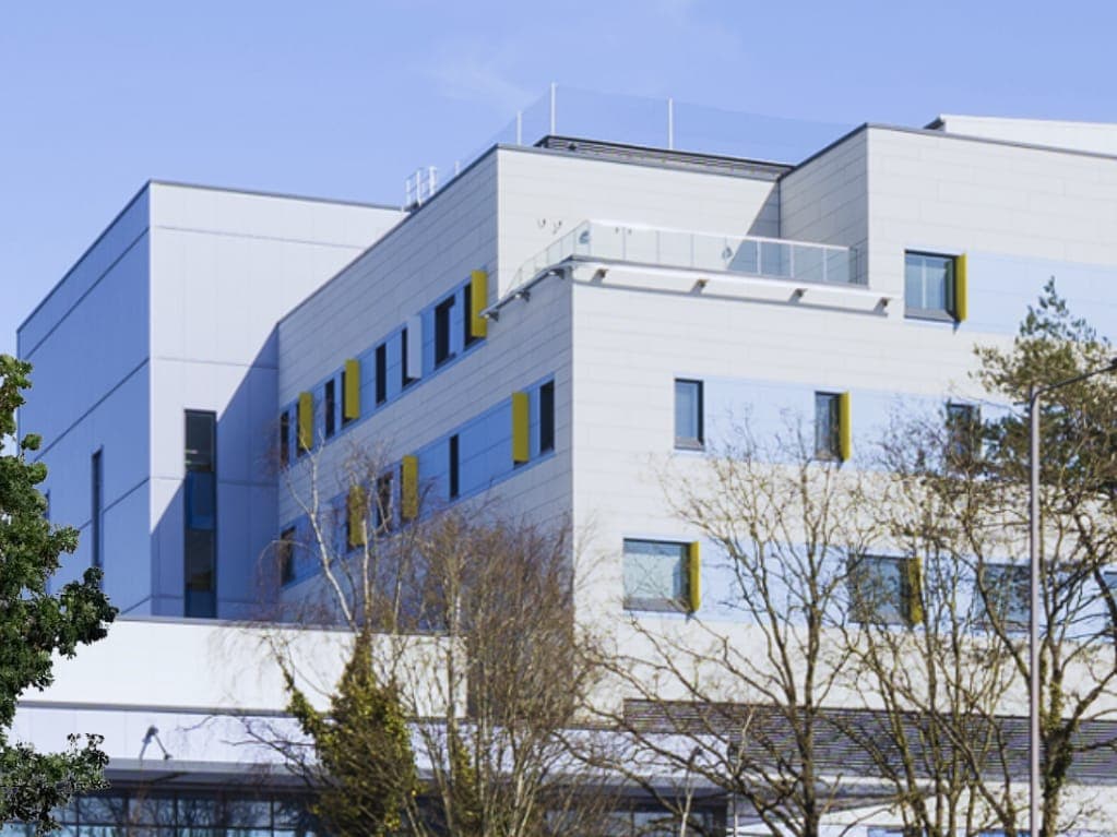 A modern multi-storey building with pale cladding and trees in front of it, under a blue sky