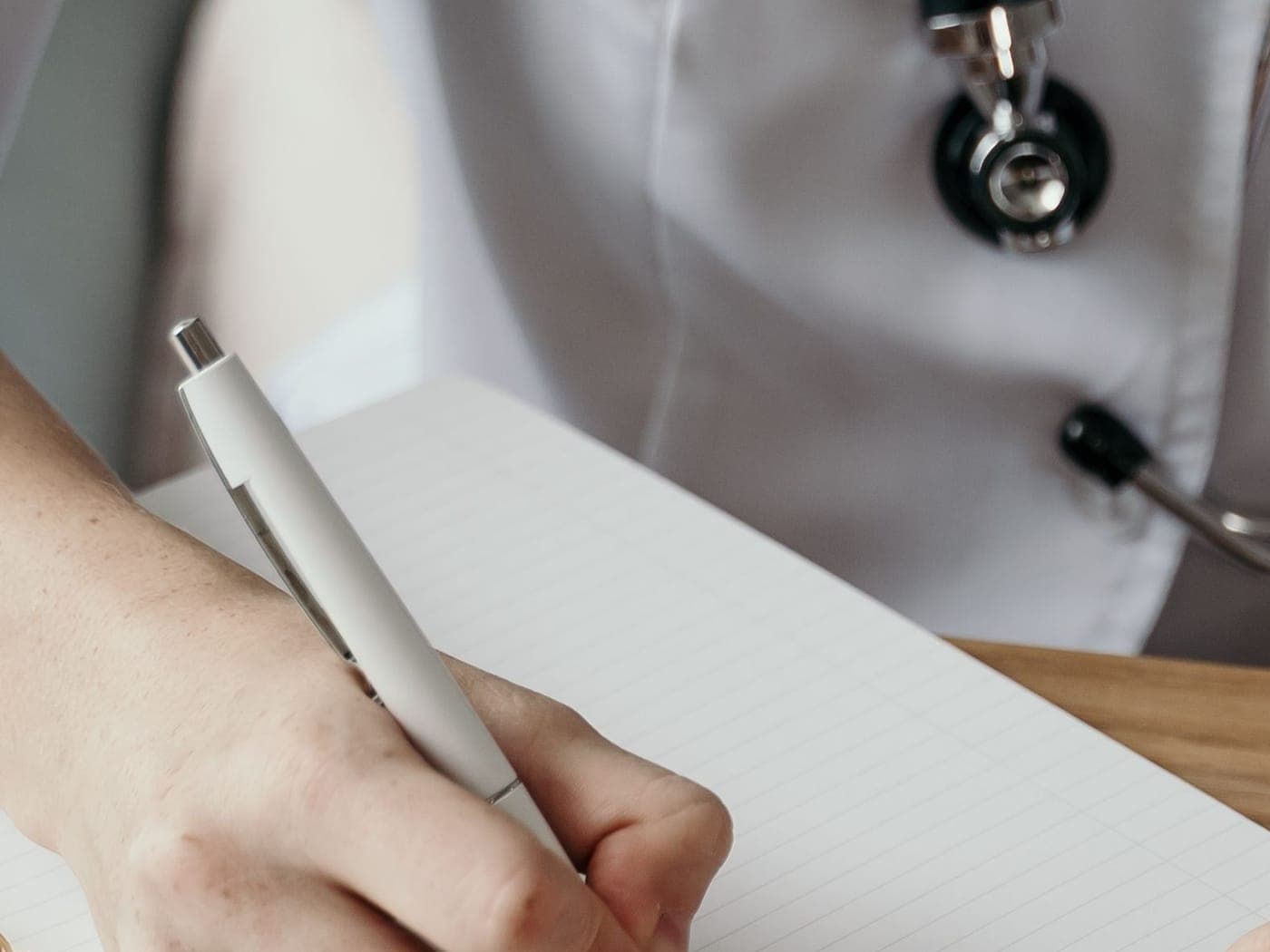 A person in a white coat with a stethoscope writing in a notebook at a wooden desk