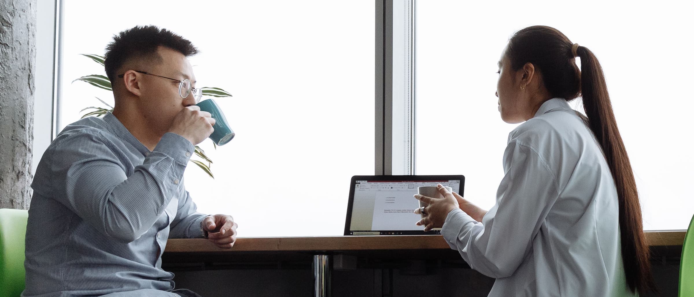 Two people seated at a table by a large window, one drinking from a mug and one in a white coat in front of a laptop
