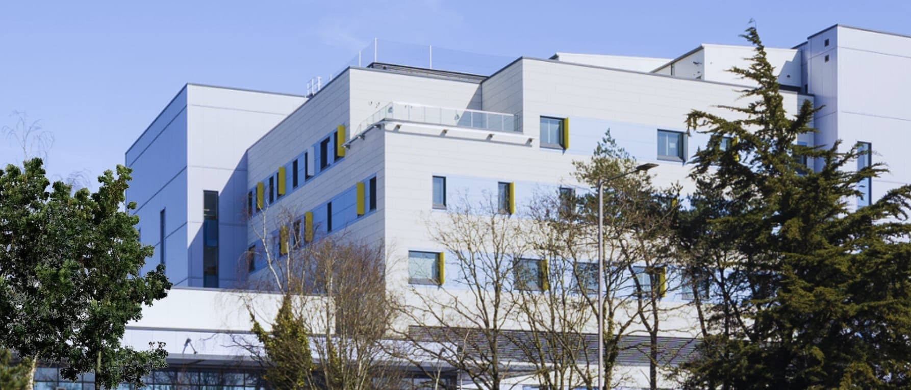 A modern multi-storey building with pale cladding and trees in front of it, under a blue sky