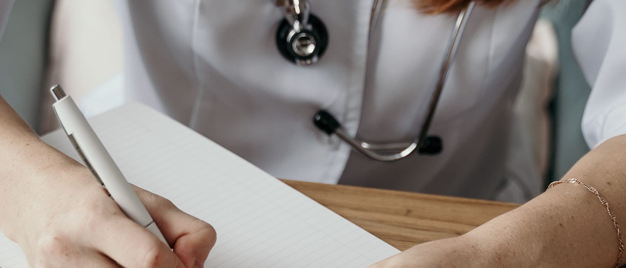 A person in a white coat with a stethoscope writing in a notebook at a wooden desk